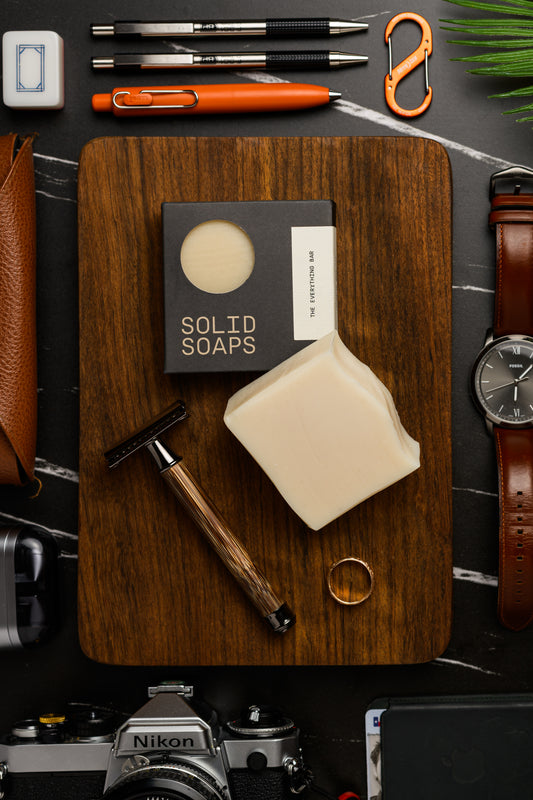 Solid Soaps The Everything Bar on a wooden board with a razor, camera, and other items on a dark surface